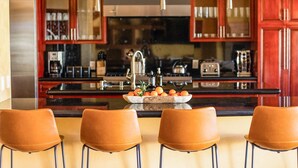 A kitchen with wooden cabinets, a black countertop, and a central island with a bowl of oranges. There are three brown chairs in front of the island. - Amarilla by Avantstay Peaceful Paradise w/ Pool, Spa & Theatre Room (La Quinta)