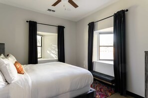A hotel room with a large bed, two windows with black curtains, a ceiling fan, and a colorful rug.