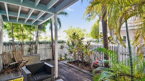Un área de patio techada con muebles de mimbre, cerca blanca de piquete y plantas tropicales.