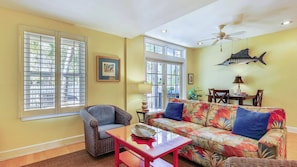 A living room with a sofa, chair, and table, large windows, and a ceiling fan. - Key West Casa by Avantstay Communal Pool BBQ & Patio Near Duval Street Month Long Stays Only (Key West)