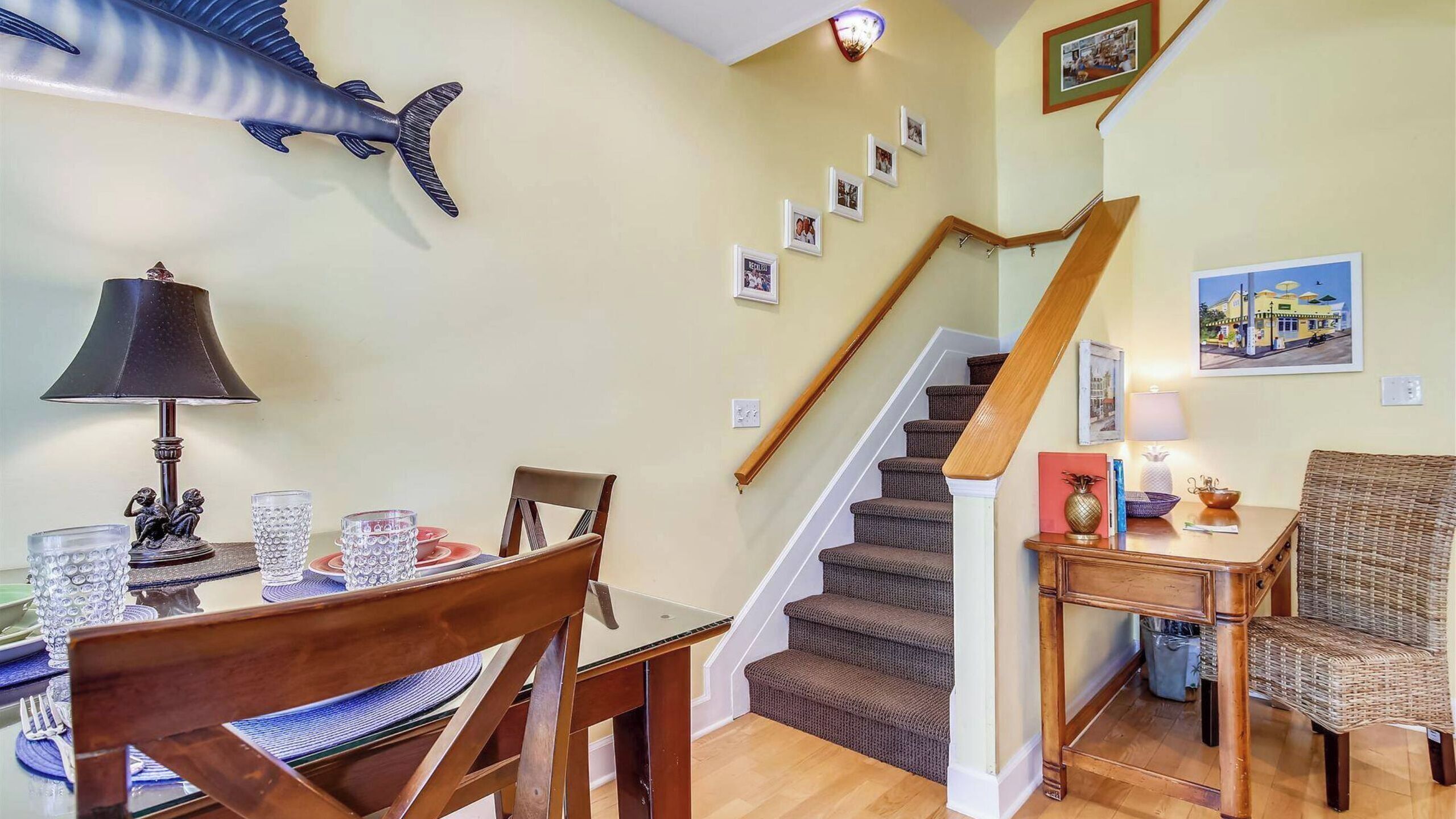 Un comedor con una mesa, sillas y una lámpara. Se ve una escalera con barandilla de madera y peldaños alfombrados.