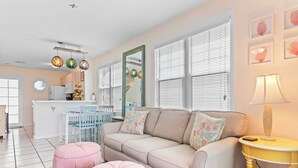 A living room with a beige sofa, pink ottomans, a yellow side table, and a white dining area with blue chairs. - Coral Breeze by Avantstay Close to Beach w/ Balcony & Shared Pool! Month Long Stays Only (Key West)