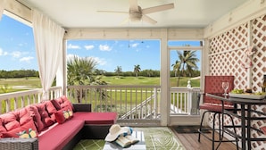 A balcony with a red sofa, a table with a glass, and a view of a golf course. - Coral Breeze by Avantstay Close to Beach w/ Balcony & Shared Pool! Month Long Stays Only (Key West)