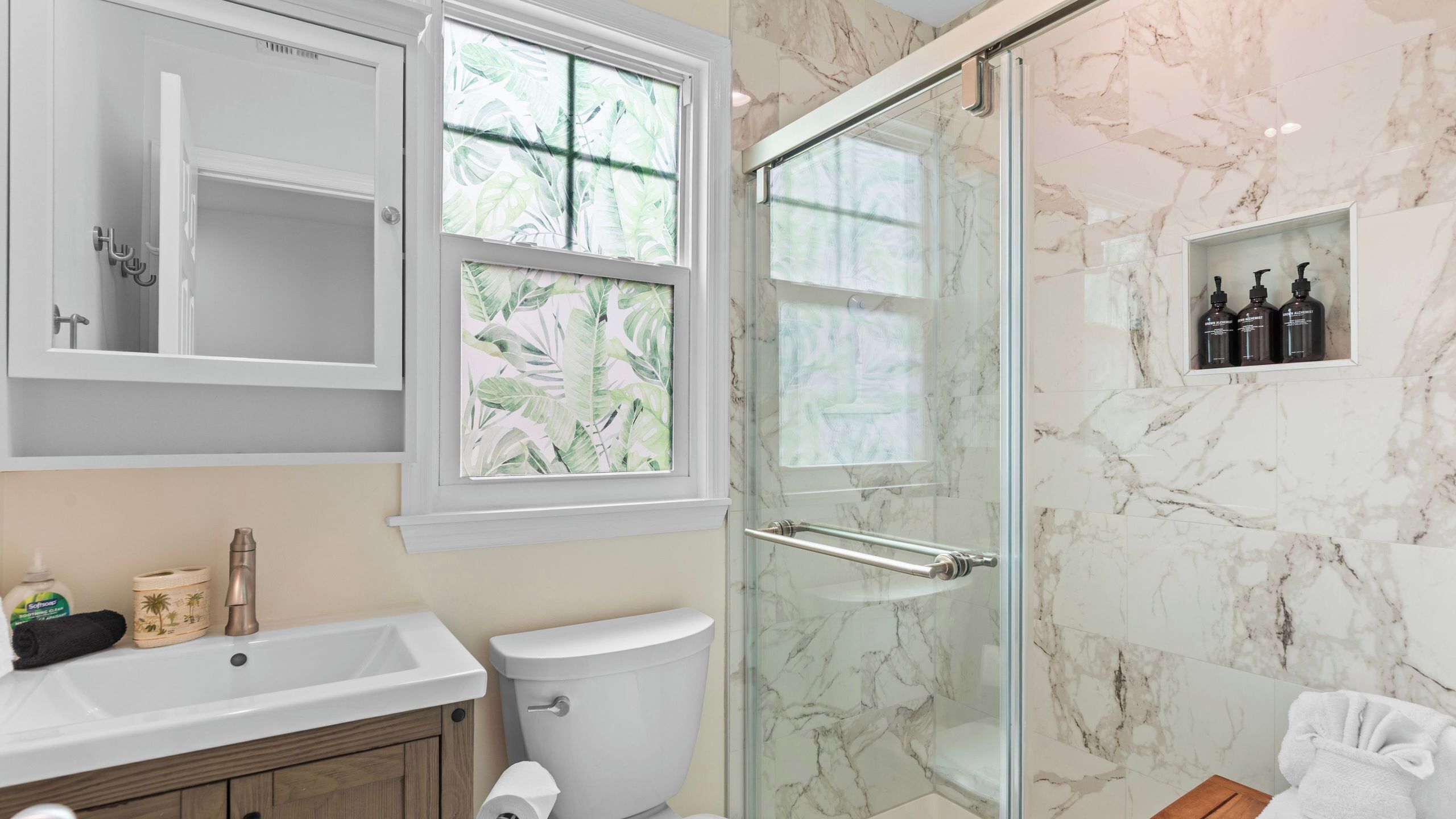 A bathroom with a shower, toilet, sink, and a window with a tropical leaf pattern.