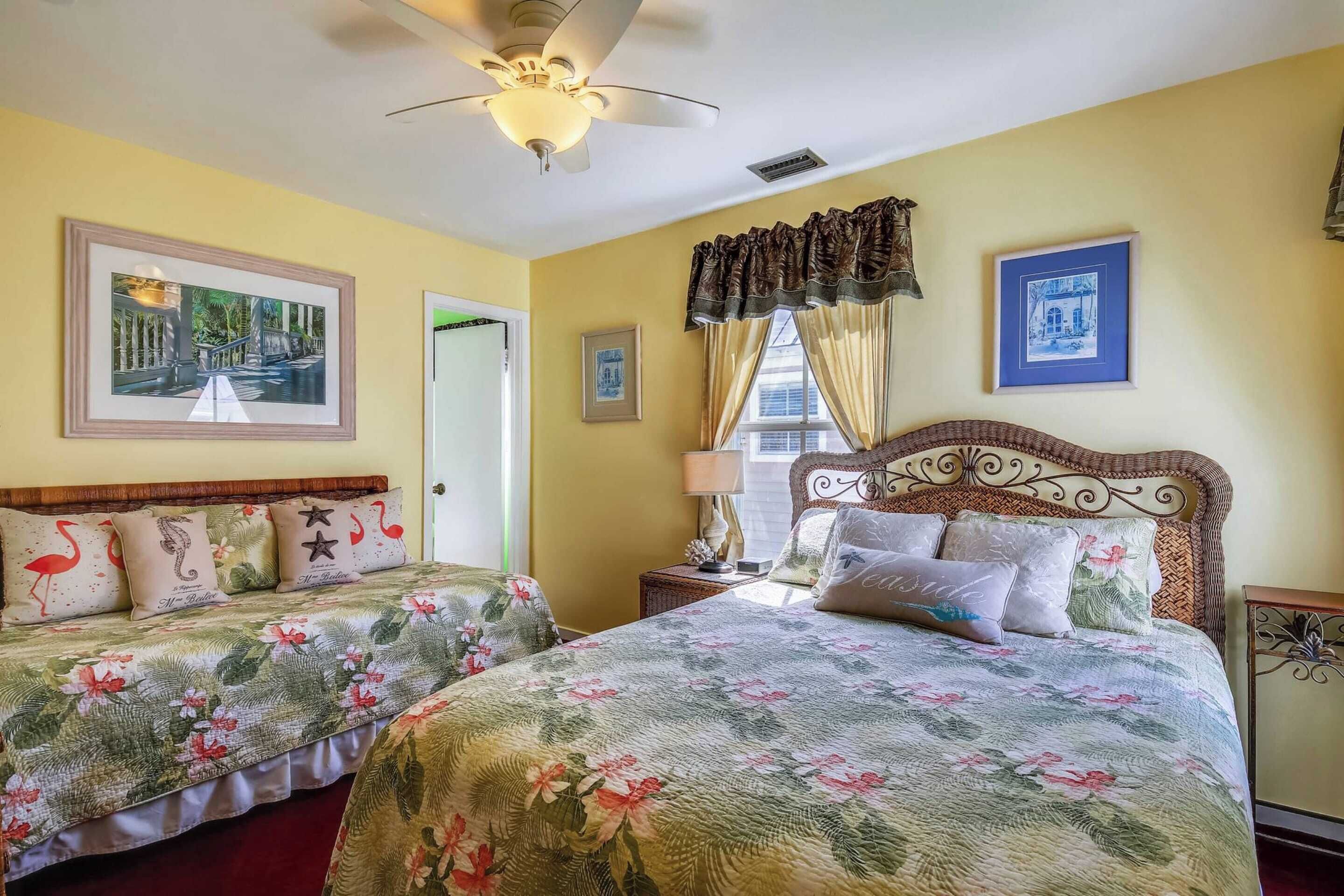 A hotel room with two beds, floral bedding, a ceiling fan, and artwork on the walls.