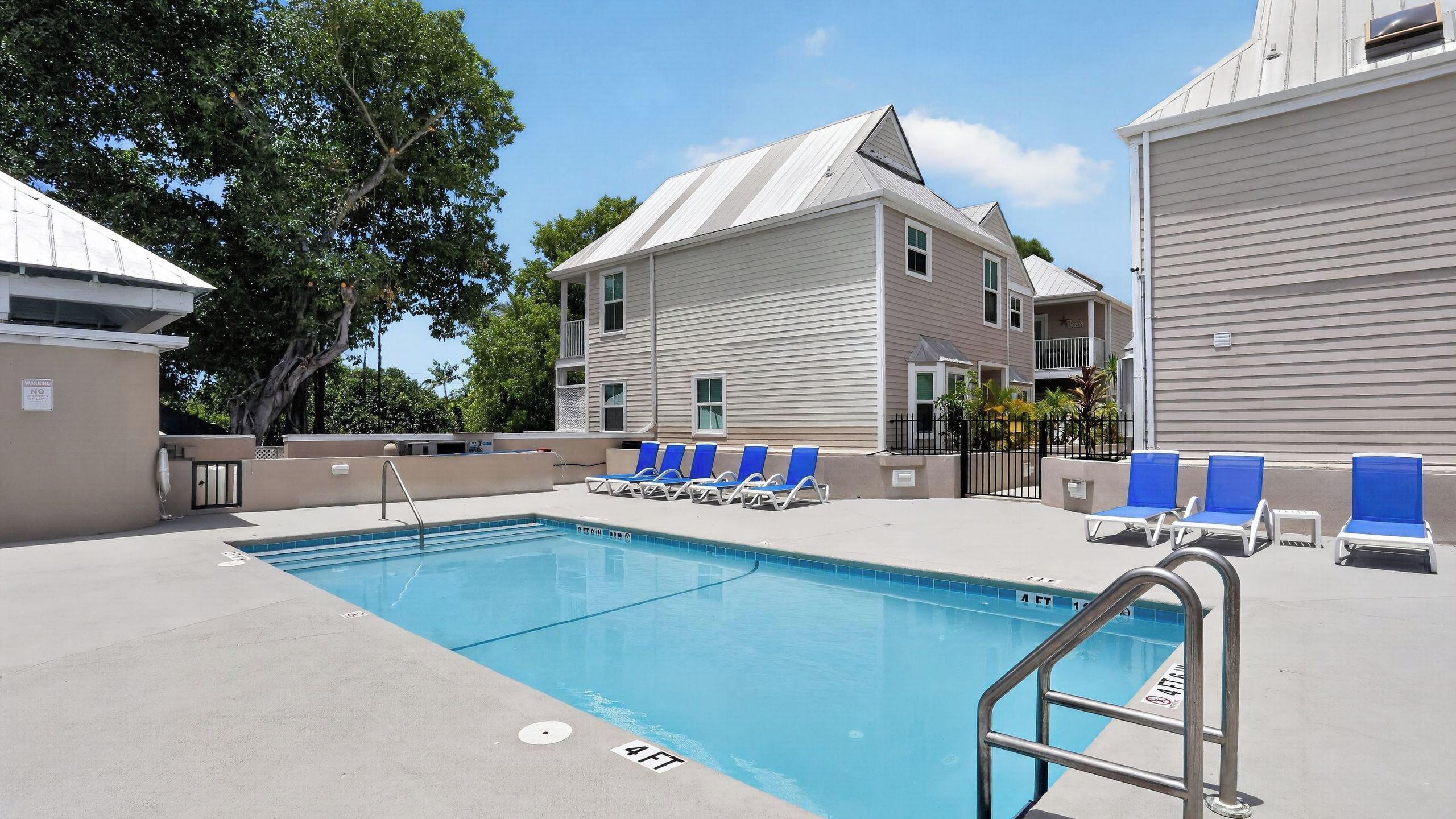 A swimming pool with blue chairs, a metal ladder, and a building with beige siding and a sloped roof.