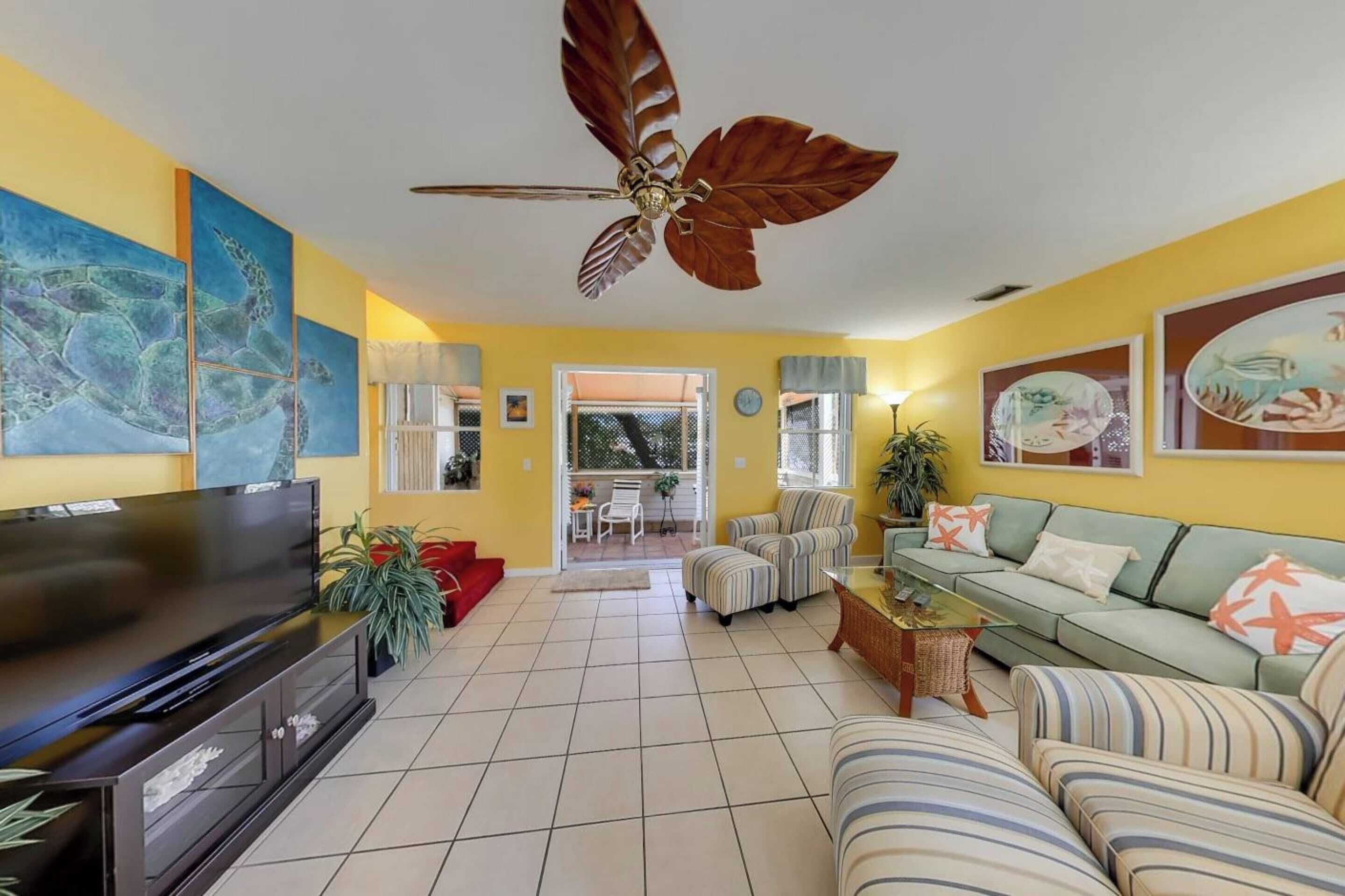 A living room with a large flat-screen TV, a ceiling fan, striped sofas, and a dining area with a table and chairs.