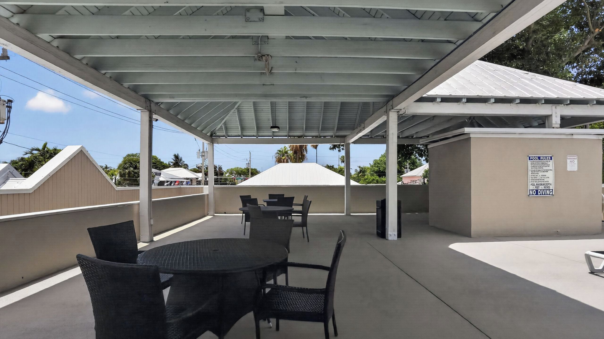 An outdoor covered patio with tables and chairs, a pool with a sign indicating no diving, and a building with a white roof in the background.