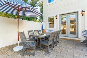 Un patio exterior con una mesa lista para comer, rodeado de sillas y una sombrilla a rayas que da sombra.
