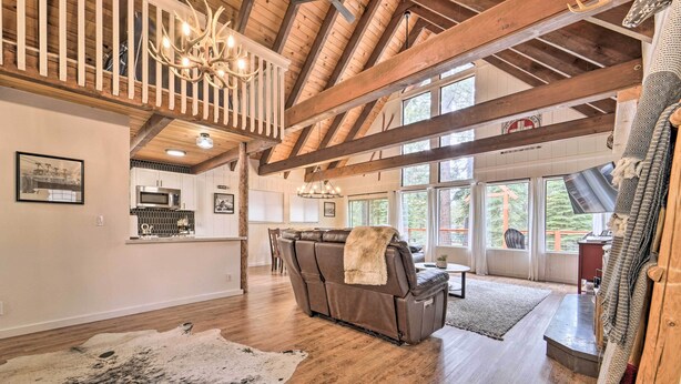A spacious living room with a leather sofa, a chandelier, and a large window offering a view of the outdoors. - 2 Mi to Tahoe Donner Ski Resort: Forest-View Haven (Truckee)
