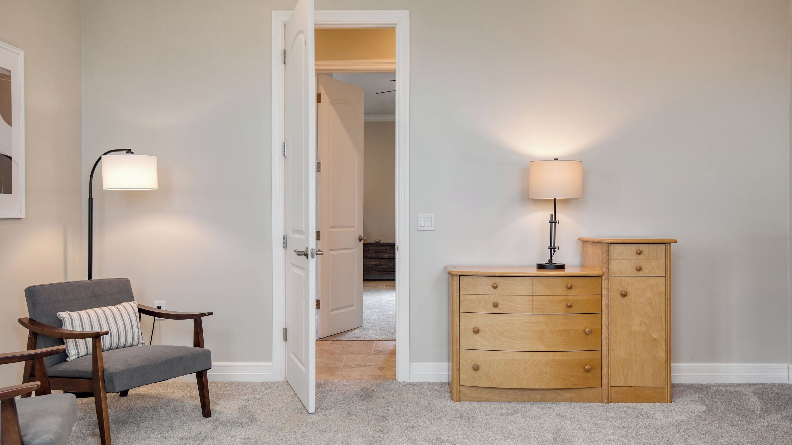 Una habitación con un sillón gris, un aparador de madera, una lámpara de pie y una puerta que da a otra habitación.