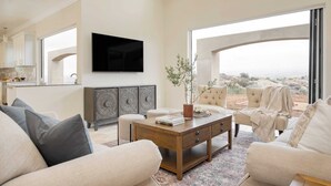 A spacious living room with a large window, a flat-screen TV mounted on the wall, and a coffee table with books and a plant.