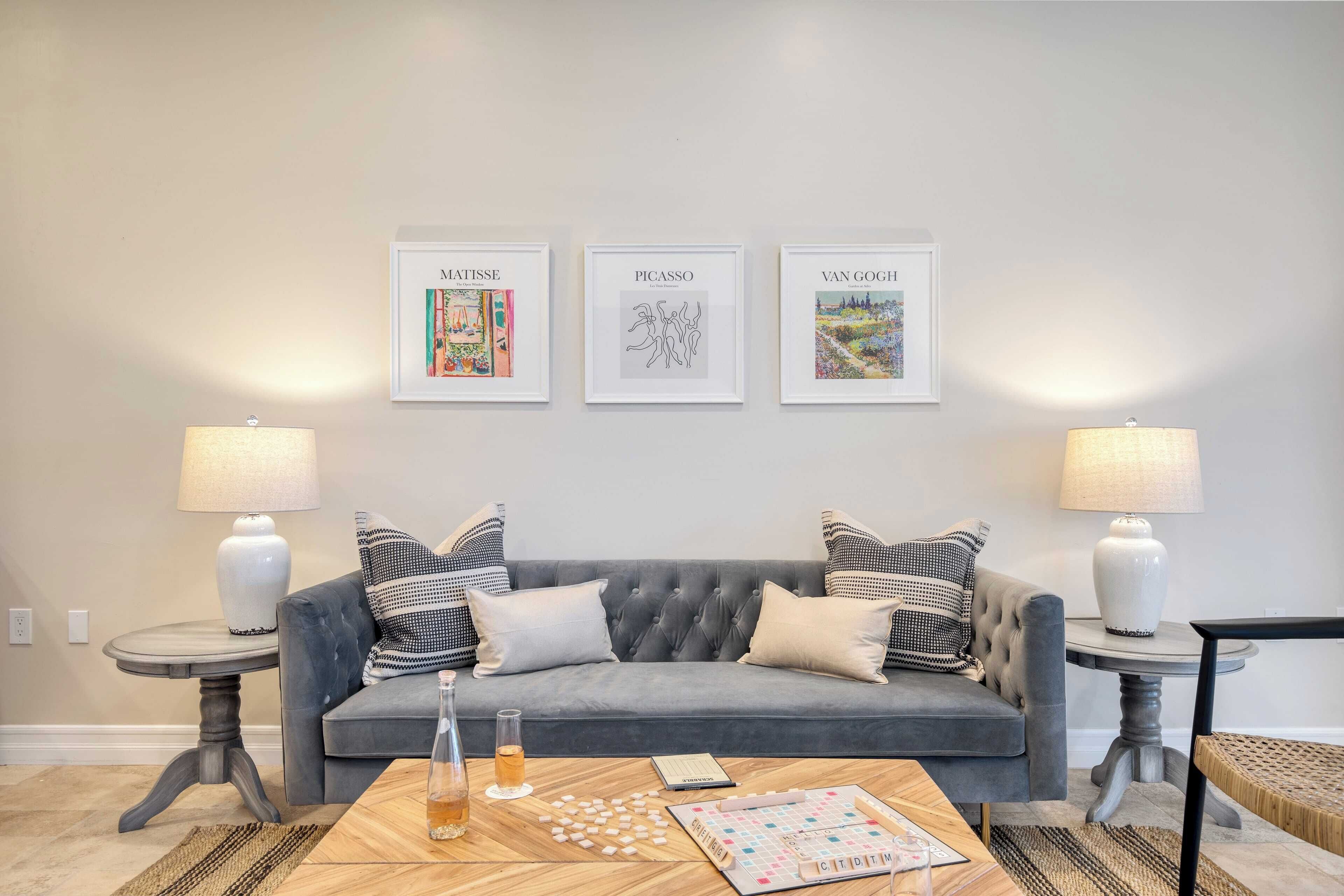 A living room with a grey sofa, two side tables with lamps, a coffee table with a board game, and three framed artworks on the wall.