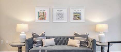 A living room with a grey sofa, two side tables with lamps, a coffee table with a board game, and three framed artworks on the wall.