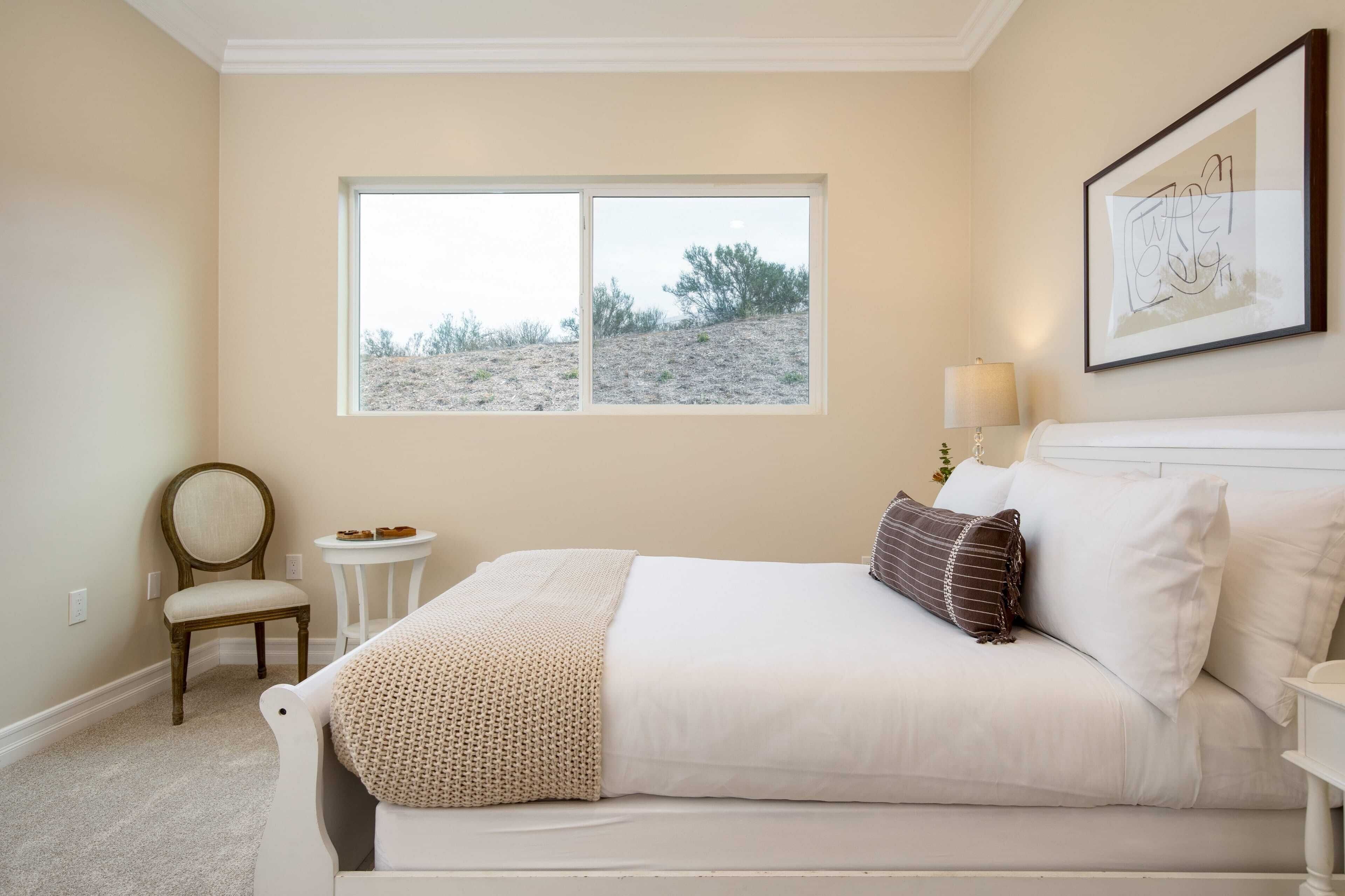 A bedroom with a bed, a chair, a small table, a window with a view of a hill, and a framed artwork on the wall.