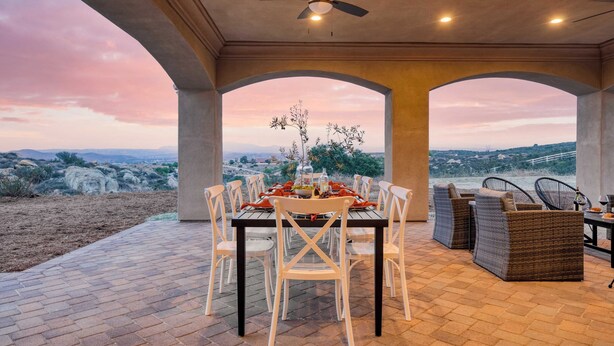 An outdoor dining area with a table set for six, surrounded by white chairs and wicker sofas, overlooking a scenic landscape.