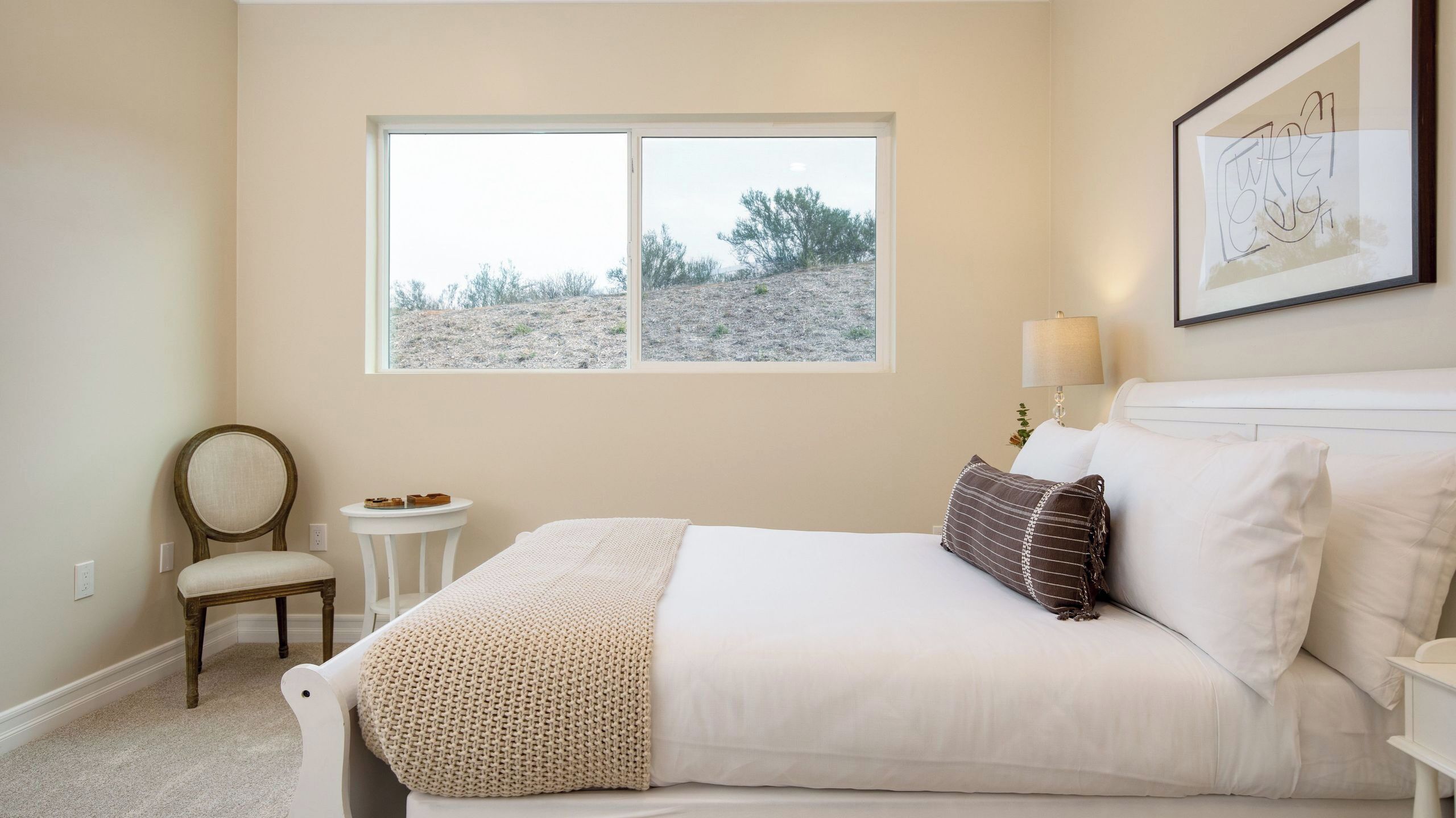 Un dormitorio con una cama, una silla, una mesita, una ventana y una lámpara de pared.