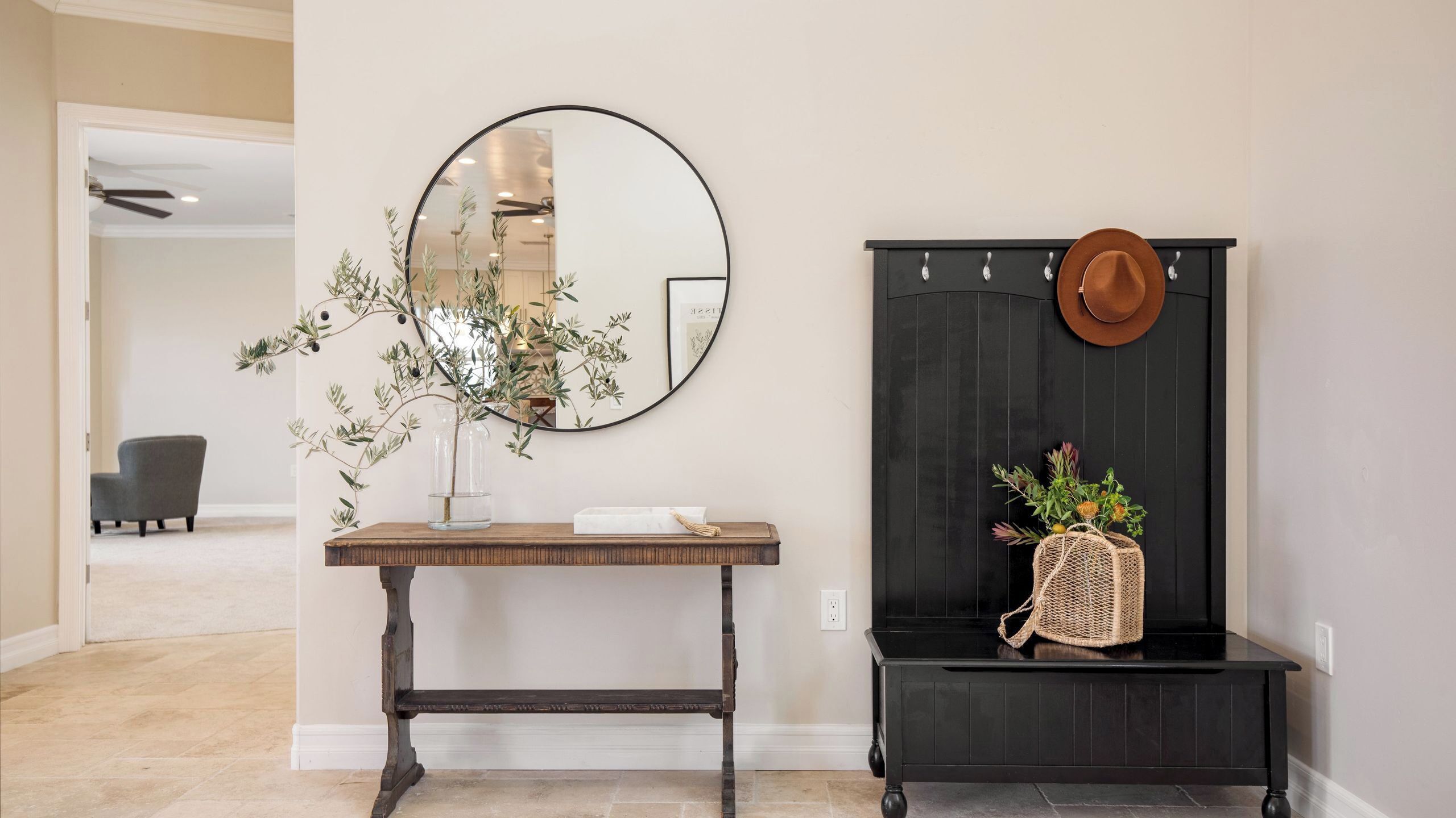 A reception area with a round mirror, a wooden console table, a bench with a woven bag, and a hat on the wall.
