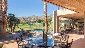 A patio with a glass table and chairs, a pool, and a view of a golf course and mountains. - Moonshine by Avantstay Modern & Stylish Estate w/ Bar, Fire Pit & Hot Tub 5 bd (La Quinta)