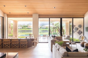 A spacious living area with a large sliding glass door leading to a poolside area, featuring a sofa, dining chairs, and a coffee table. - Moonshine by Avantstay Modern & Stylish Estate w/ Bar, Fire Pit & Hot Tub 5 bd (La Quinta)