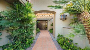 A pathway leading to a door with a wooden gate, surrounded by lush greenery and a tiled walkway.