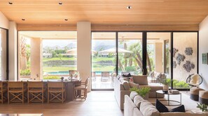 A spacious living area with a large window overlooking a pool and a golf course. - Moonshine by Avantstay Modern & Stylish Estate w/ Bar, Fire Pit & Hot Tub 5 bd (La Quinta)