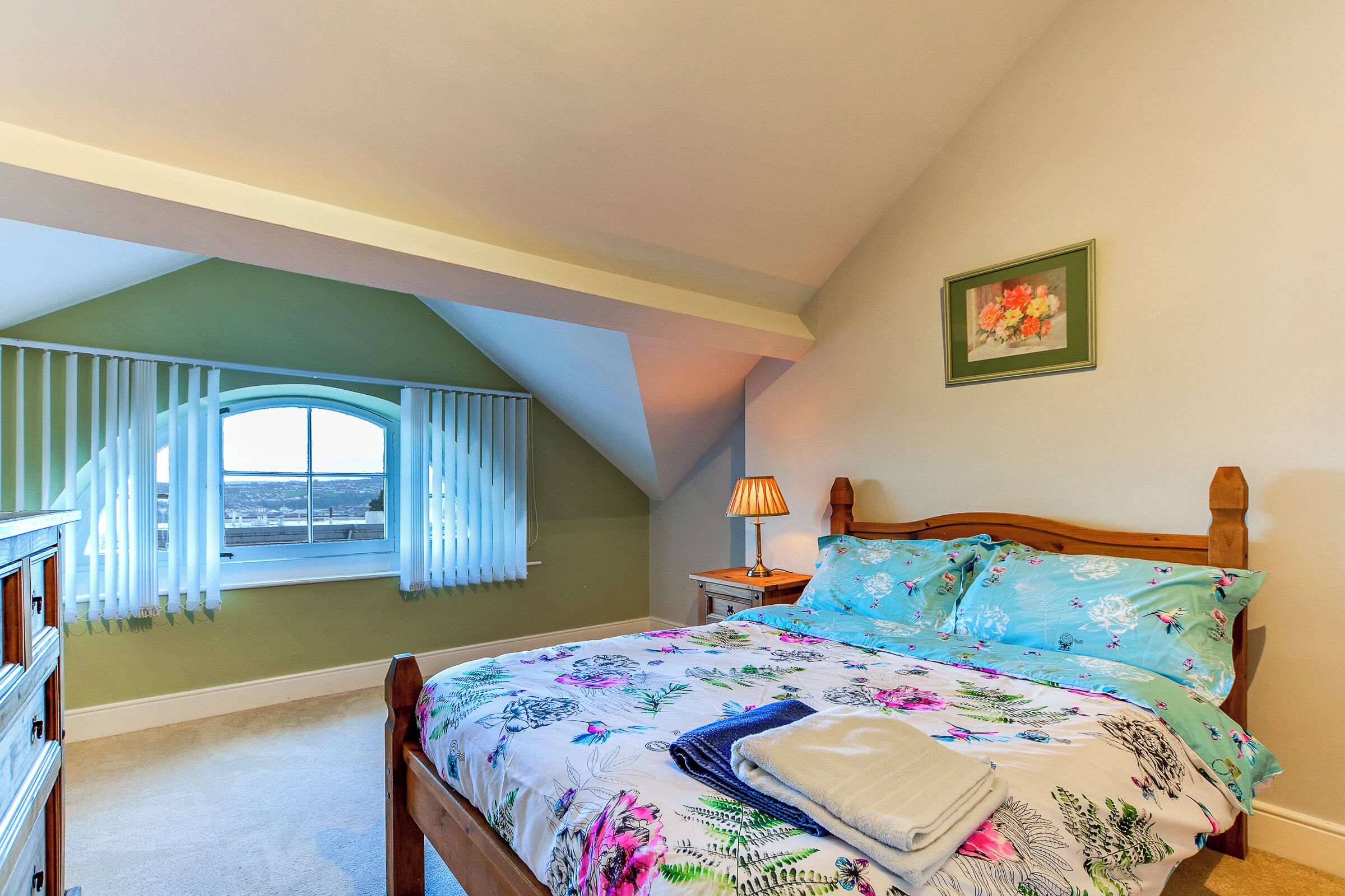 A bedroom with a sloped ceiling, a bed with floral bedding, a nightstand with a lamp, and a window with curtains.