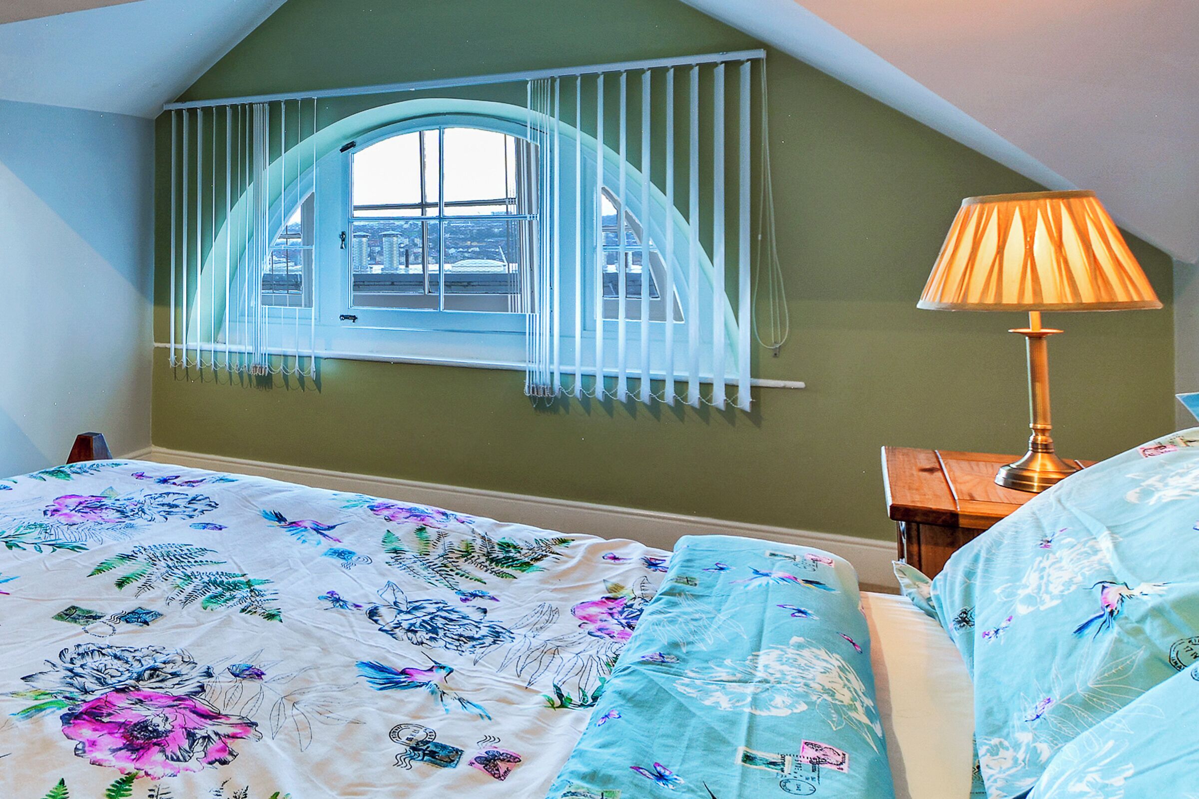 A bedroom with a bed, a window with curtains, a lamp, and a view of the city.
