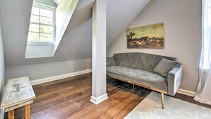 Una habitación con un sofá gris, un pilar blanco, un cuadro de un paisaje y una ventana con vistas a árboles.