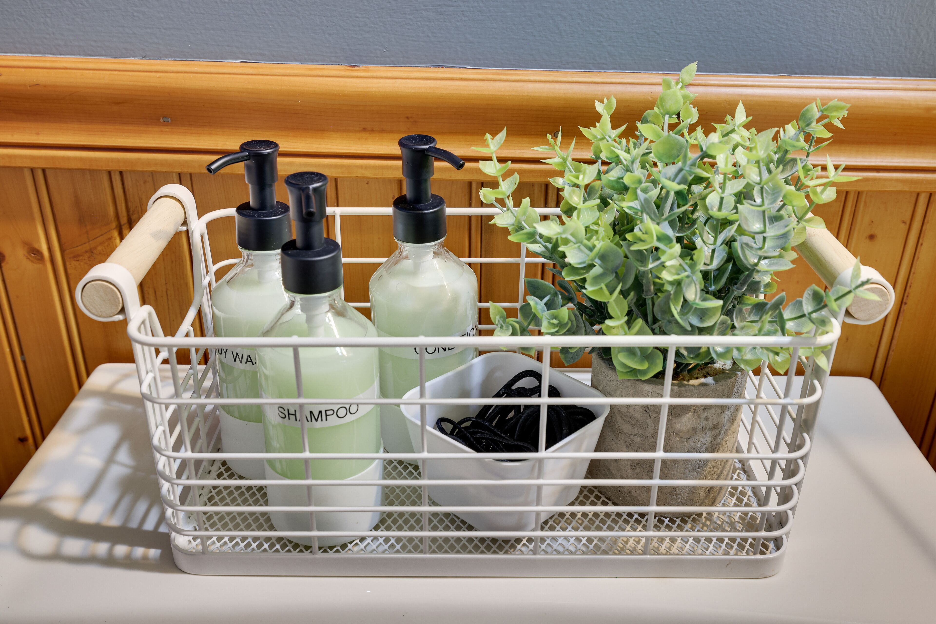 Una cesta metálica blanca con botellas etiquetadas como 'SHAMPOO' y 'COOLING', una pequeña planta en maceta y un recipiente blanco con objetos negros.