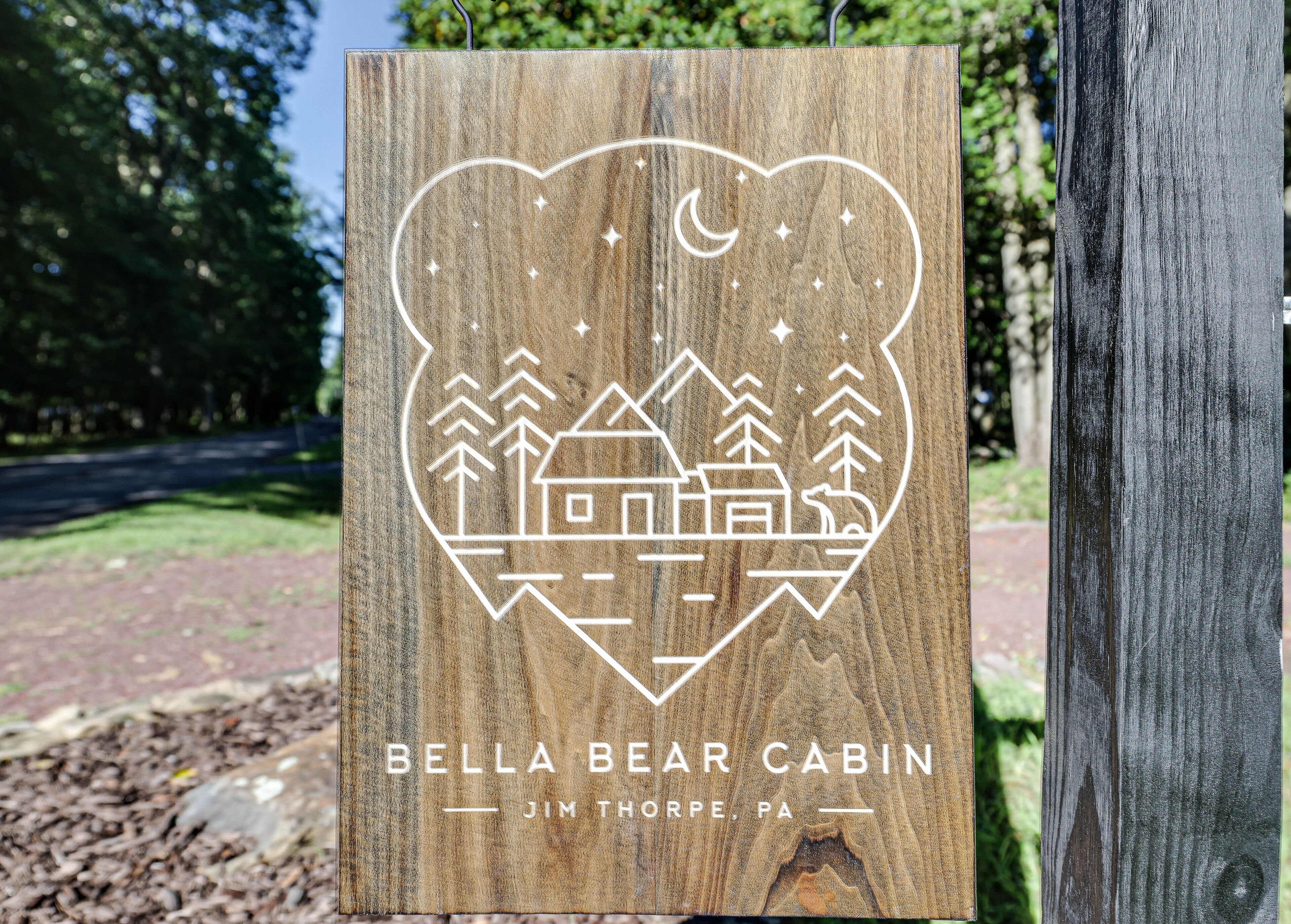 A wooden sign with a heart-shaped cutout featuring a cabin, trees, and a moon. Text reads "Bella Bear Cabin, Jim Thorpe, PA".