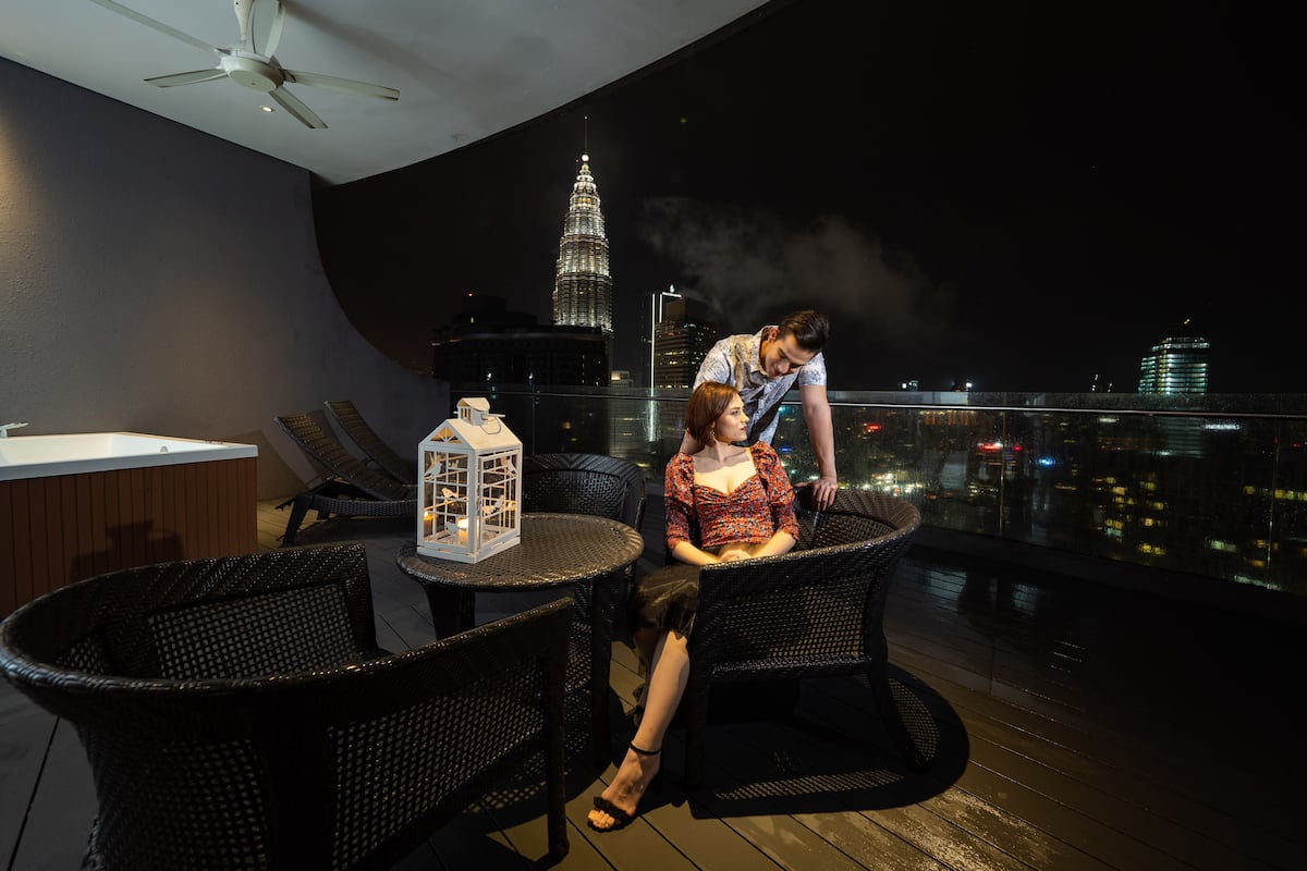 A balcony with wicker chairs, a small table with a lantern, and a view of a city skyline at night.