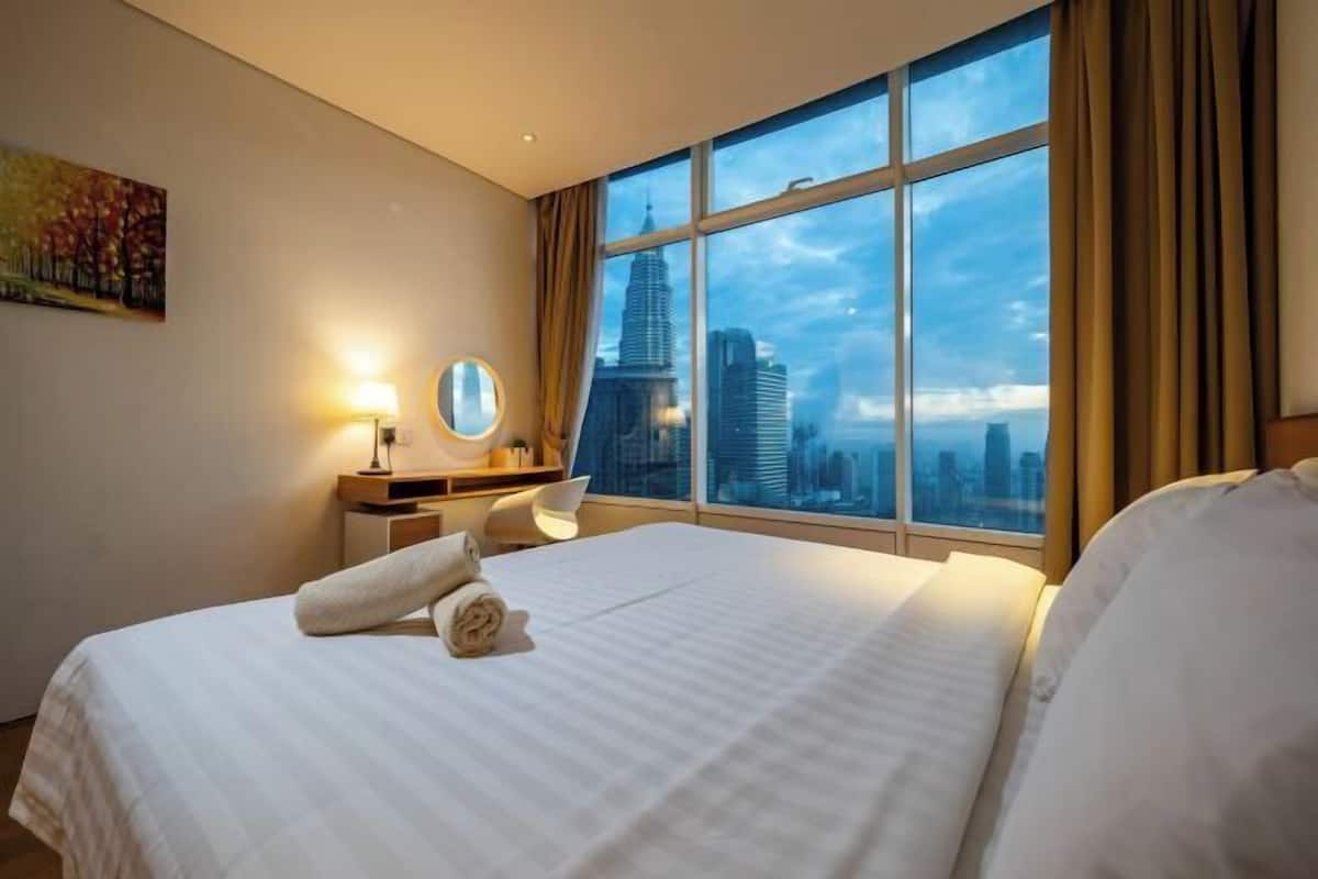 A hotel room with a large bed, a bedside table with a lamp, a round mirror, and a view of a cityscape through a large window.