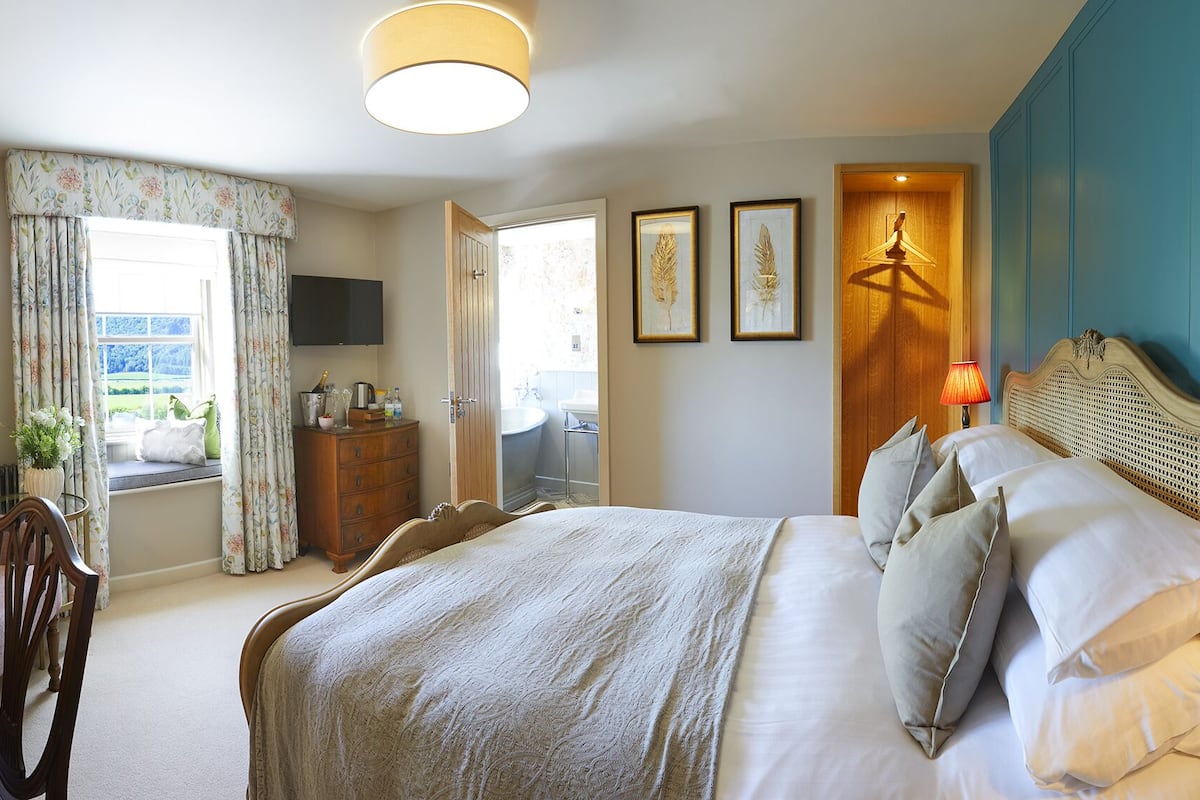 Un dormitorio con cama, mesita de noche, lámpara, ventana con cortinas y vistas al exterior.