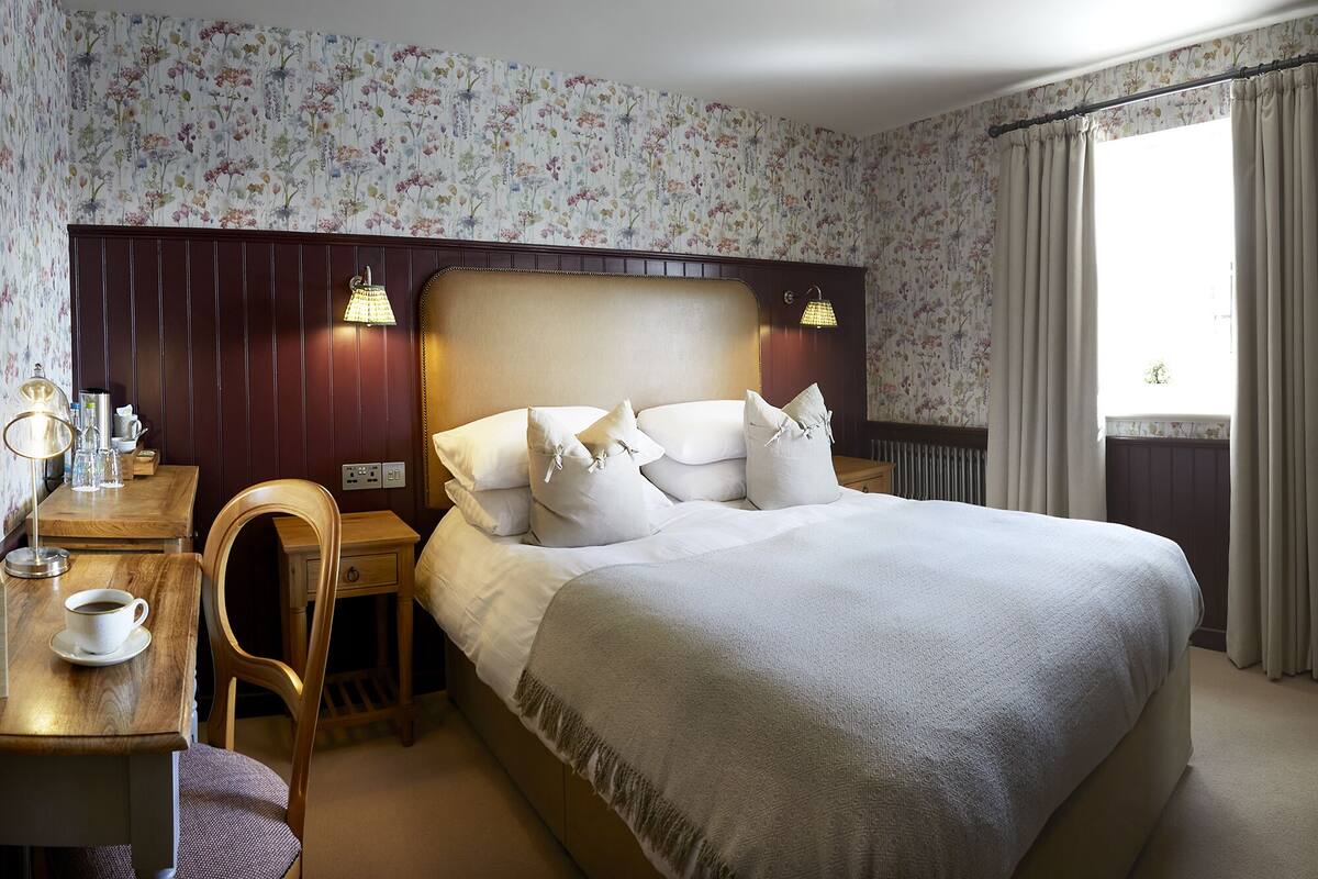 Habitación de hotel ordenada con una cama grande, una mesita de noche de madera con una lámpara, una silla y una mesa de centro.
