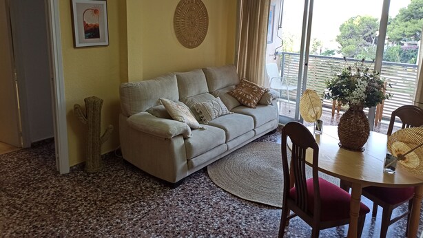Una sala de estar con un sofá, una mesa de comedor con sillas, un ventanal con cortinas, un adorno en la pared y una planta de cactus.