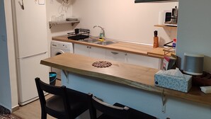 Una cocina compacta equipada con refrigerador, lavadora y cafetera. Hay dos sillas y una mesa con camino de mesa.