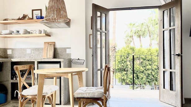 A kitchen with a dining area, wooden chairs, a hanging light fixture, and a view of greenery through open doors. - Loreto80– Studio Palo Blanco (Loreto)