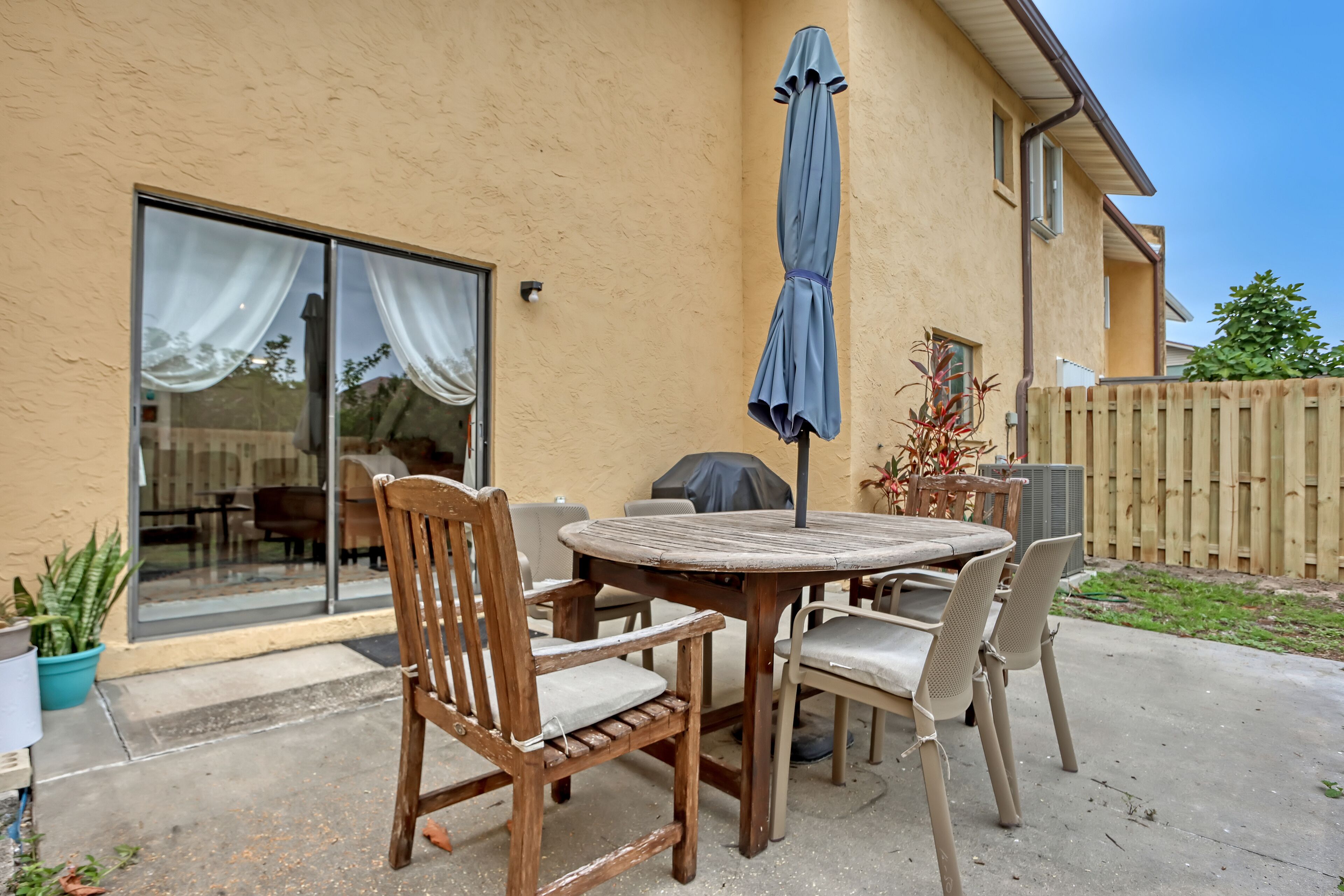 A patio with a round table and chairs, a blue umbrella, and a sliding glass door leading to an indoor area.