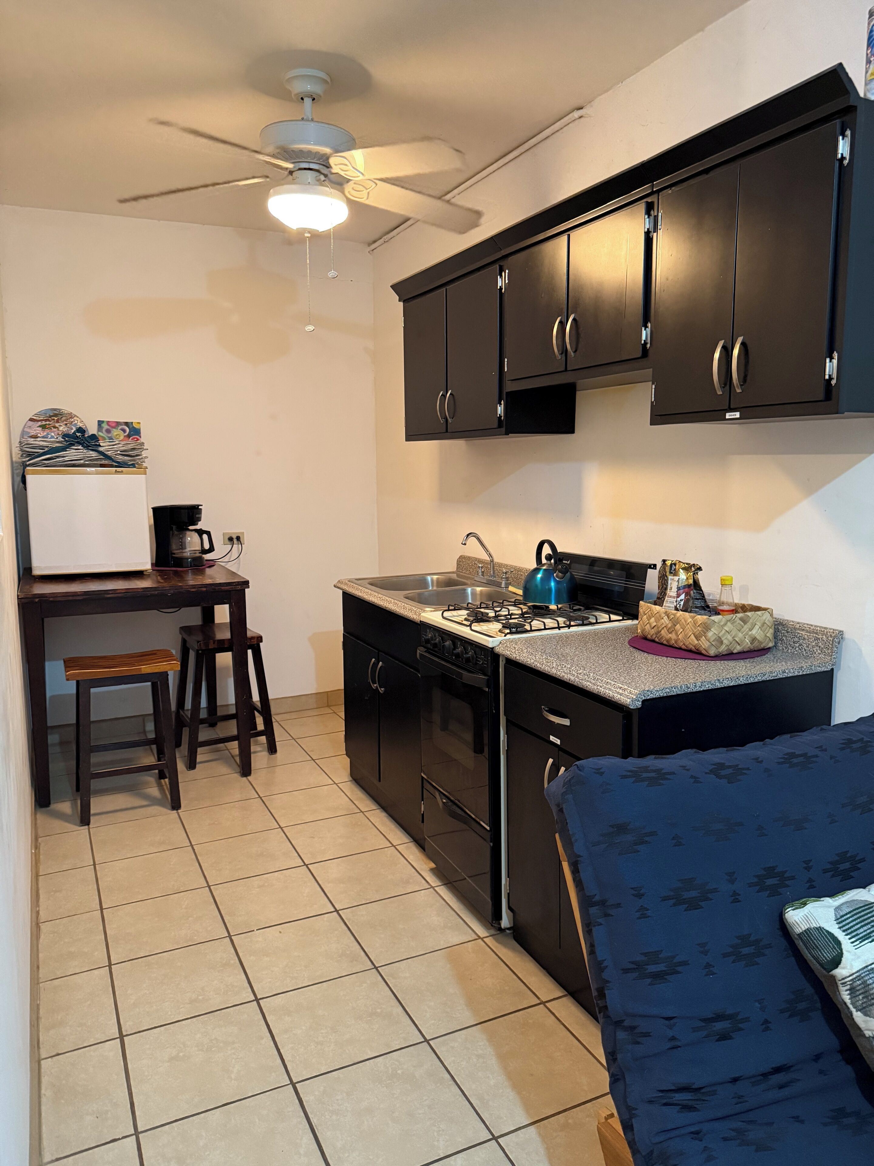 A compact kitchen with dark cabinets, a gas stove, a sink, and a ceiling fan. There is a small dining area with stools and a refrigerator.