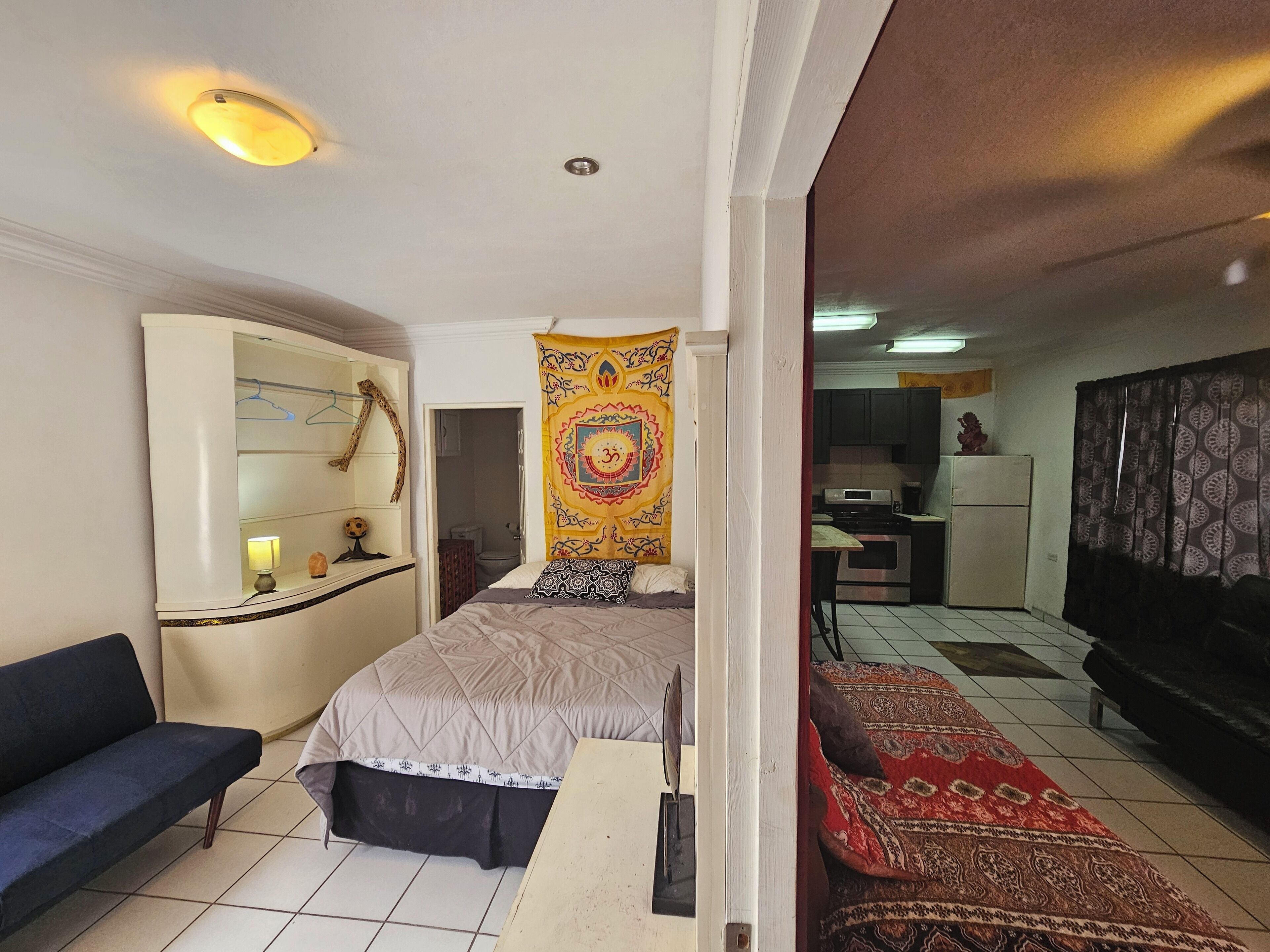 Ein Schlafzimmer mit Bett, Nachttisch, Lampe, Spiegel, Kleiderschrank, Wandteppich, einer Couch und einem durch eine offene Tür einsehbaren Küchenbereich.