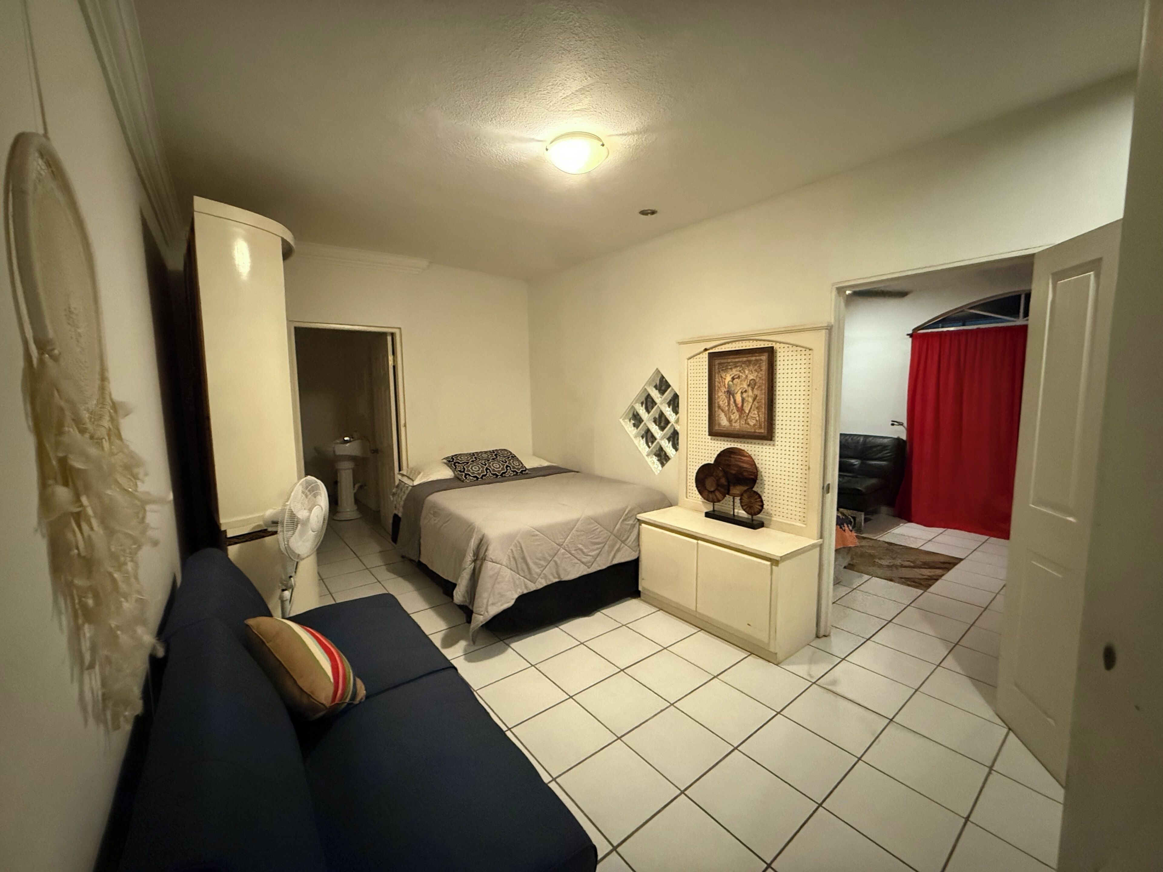 A bedroom with a bed, a bedside table, a dresser, a fan, and a red curtain.