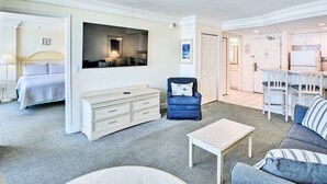 Une chambre d’hôtel comprenant un lit, une commode, une chaise, un canapé, une table et un coin cuisine visible à travers une porte ouverte.