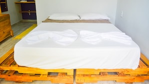 Un lit simple posé sur une palette en bois, avec une literie blanche, une commode en bois et une étagère avec des bols.