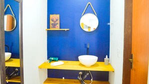 Une salle de bain avec un mur bleu, une étagère en bois, un lavabo blanc, un miroir rond, un tableau encadré et un distributeur de serviettes.