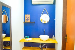Un baño con pared azul, repisa de madera, lavabo blanco, espejo redondo, cuadro enmarco y dispensador de toallas.