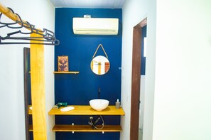 A compact bathroom with a blue wall, a white air conditioner, a wooden shelf with a white basin, a small framed picture, and a hanging round mirror.