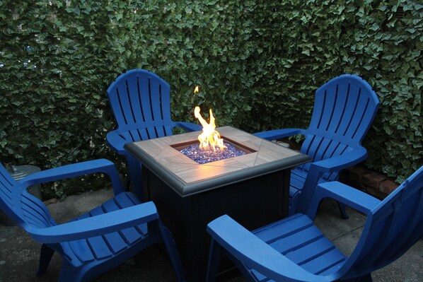 A square fire pit table surrounded by four blue chairs, set against a backdrop of dense green foliage.