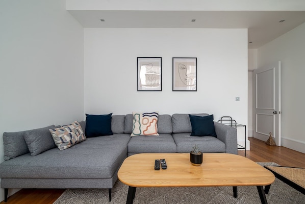 A modern living room with a grey sectional sofa, a wooden coffee table, and two framed abstract artworks on the wall. - GuestReady - Chic in the heart of Kensington (London)