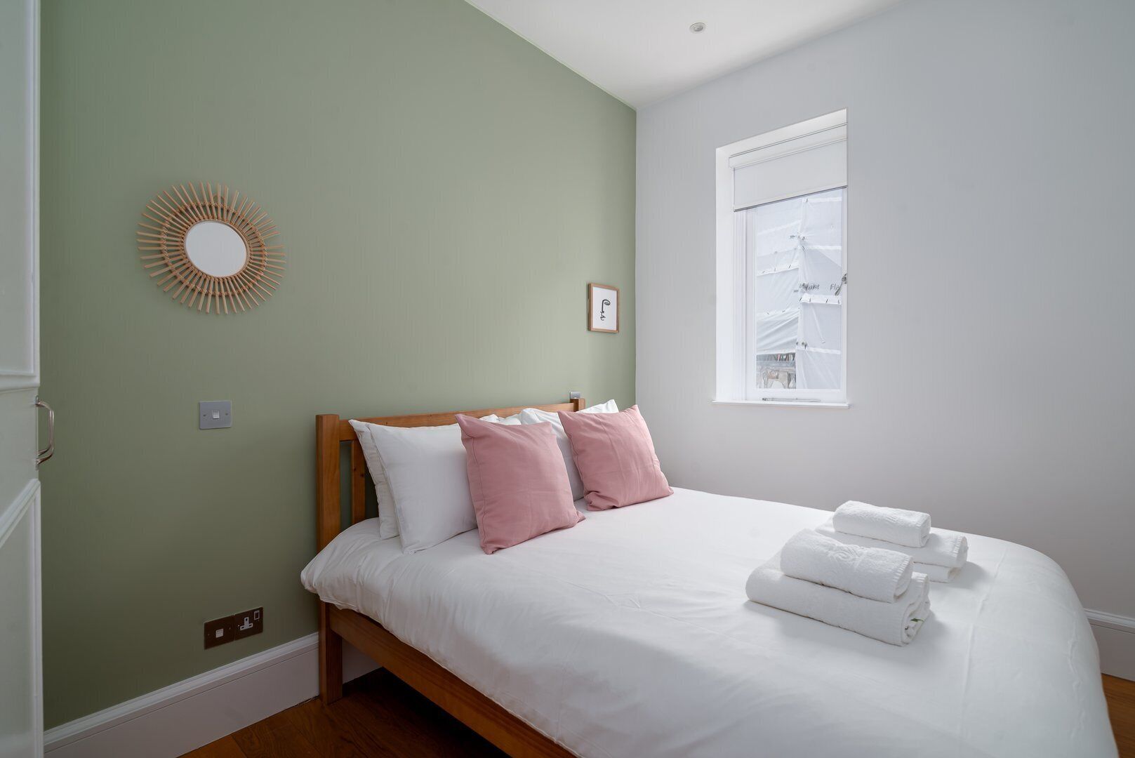 Een netjes opgemaakt bed met witte en roze kussens, een houten hoofdbord en een witte muur met een ronde spiegel en een lijst met een afbeelding.